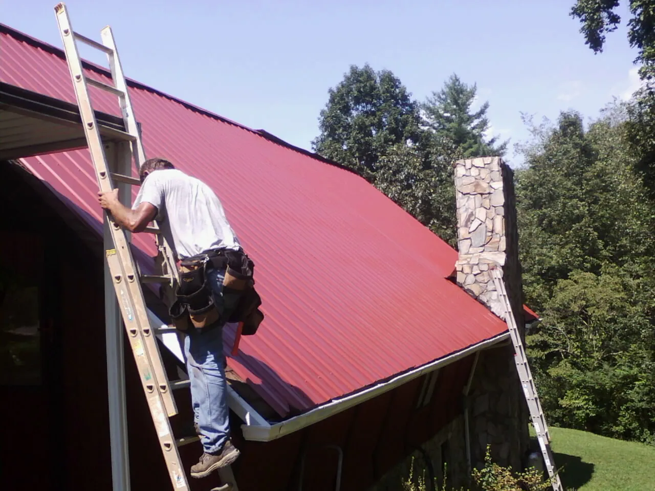 Roofer on a ladder beside a red metal roof and stone chimney — Soffit & Fascia Repair by Roofing Brothers in Rush